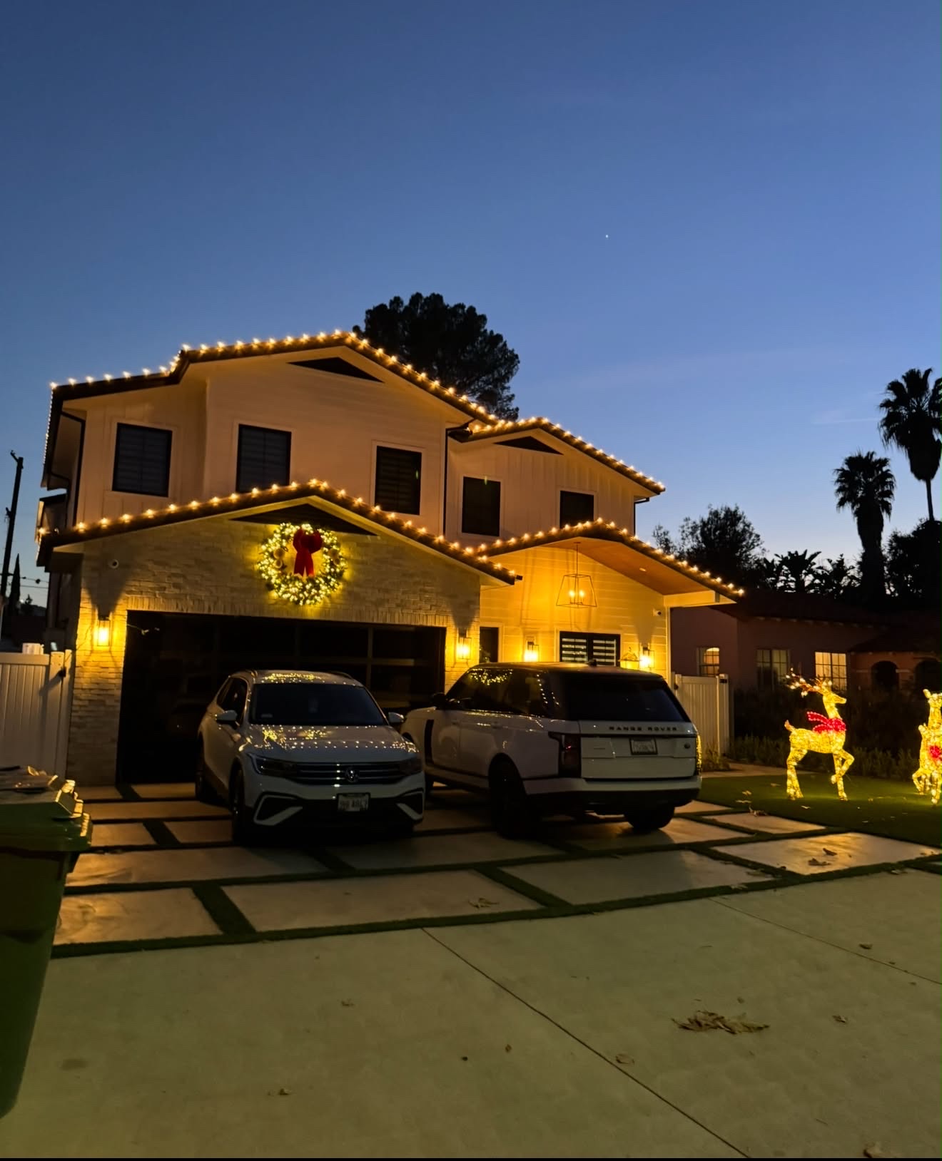 Two-story home with warm white roofline lights, lit wreath, and reindeer at dusk